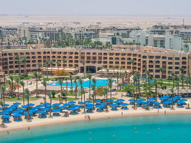 Großes Resort mit Pool, Palmen und blauen Sonnenschirmen am Strand vor klarem, türkisfarbenem Wasser.
