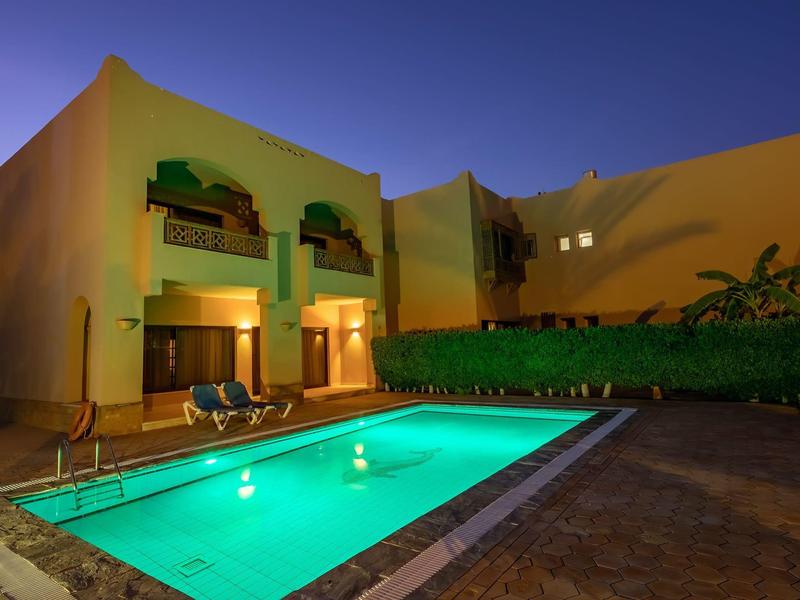 Modernes Gebäude mit gelben Wänden, beleuchtetem Pool und Sonnenschirmen bei Abenddämmerung.