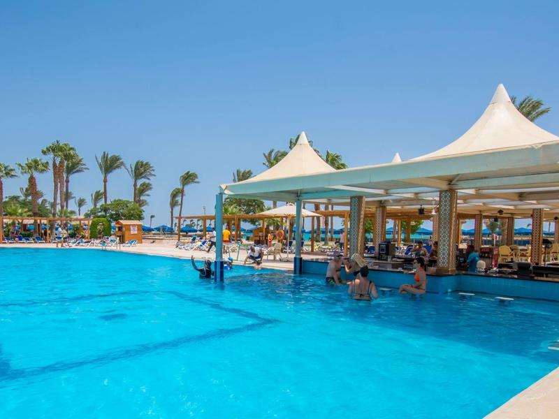 Großer blauer Pool mit barähnlichem Pavillon, Palmen und klarem Himmel im Hintergrund.