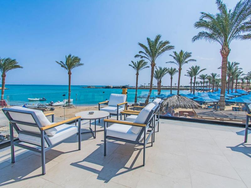 Strand mit blauen Sonnenschirmen, Palmen und modernen weißen Gartenmöbeln bei Sonne.