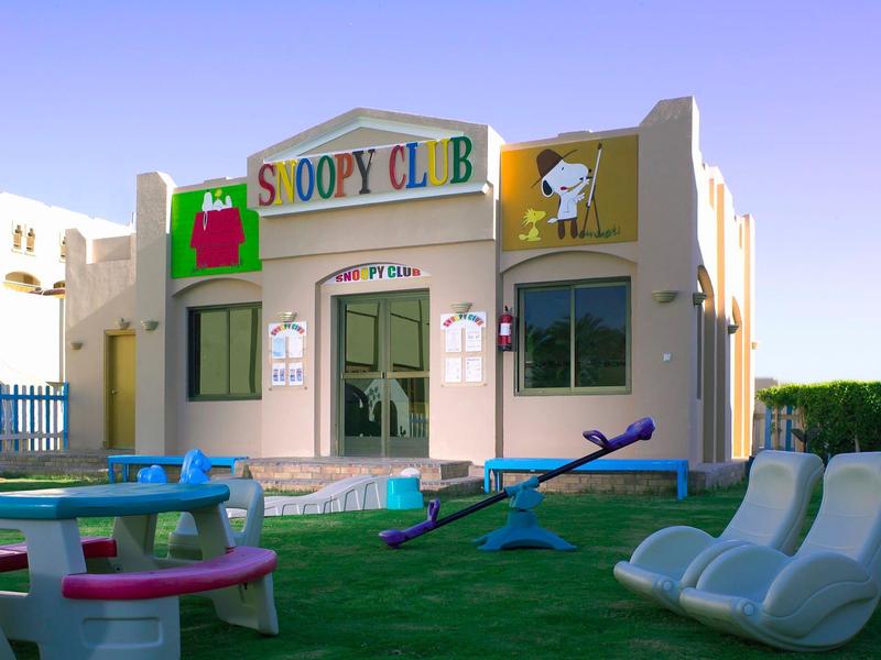 Kinderclub mit Snoopy-Motiv, grünem Rasen, Wippe, Tisch und weißen Sitzbänken im Freien.