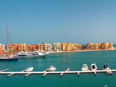 Marina avec des bateaux amarrés à une longue jetée et des bâtiments colorés au bord de la mer sous un ciel clair.