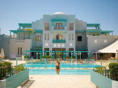 Velká bílá hotelová budova s tyrkysovými akcenty a venkovním bazénem pod jasnou oblohou.