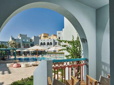 Pohled přes oblouk na bazén s lehátky a bílými budovami v pozadí.