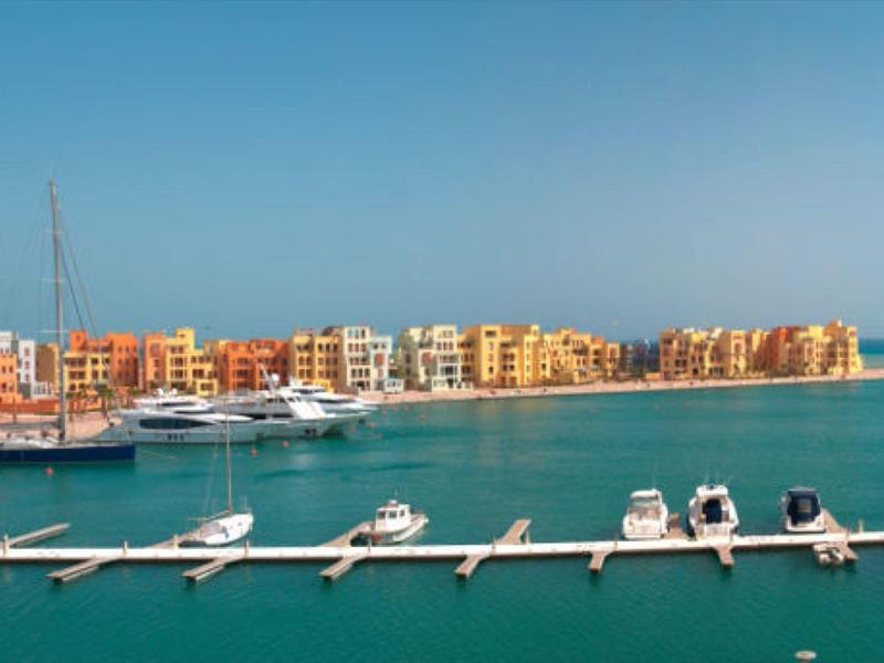Marina avec des bateaux amarrés à une longue jetée et des bâtiments colorés au bord de la mer sous un ciel clair.