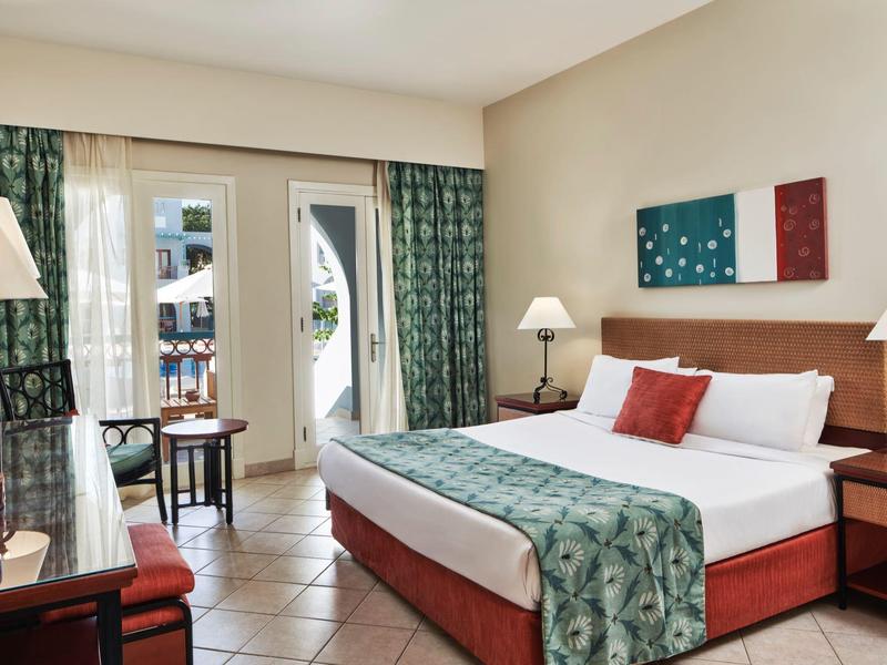 Chambre d'hôtel lumineuse avec un grand lit, des rideaux verts à motifs et un balcon extérieur.