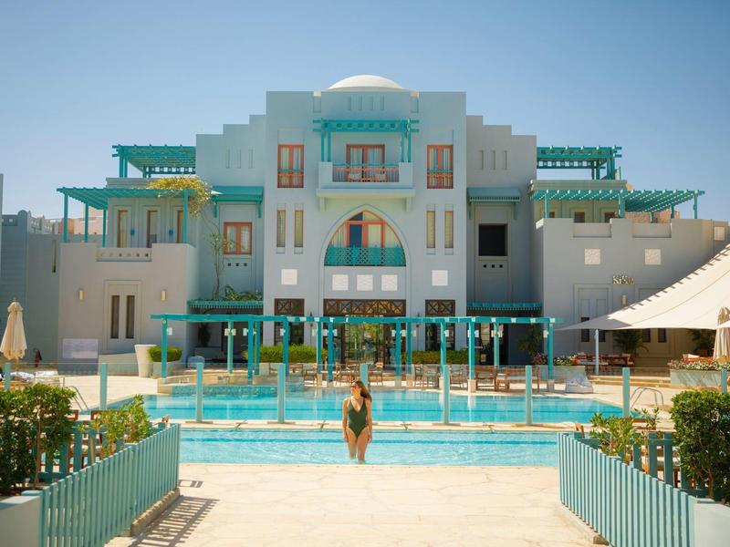 Grand hôtel blanc avec accents turquoise et piscine centrale avec une personne entrant dans l'eau.