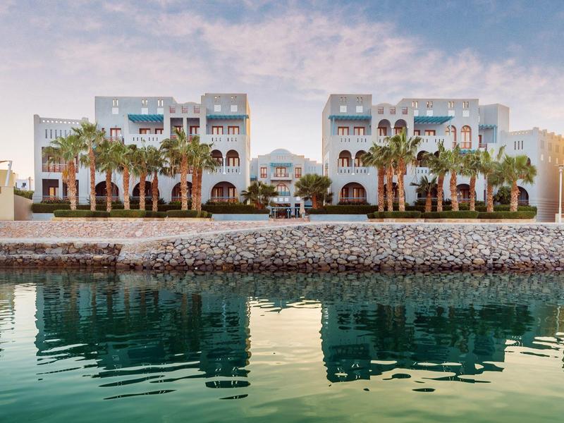 Station balnéaire avec palmiers reflétés dans une eau calme sous un ciel partiellement nuageux.