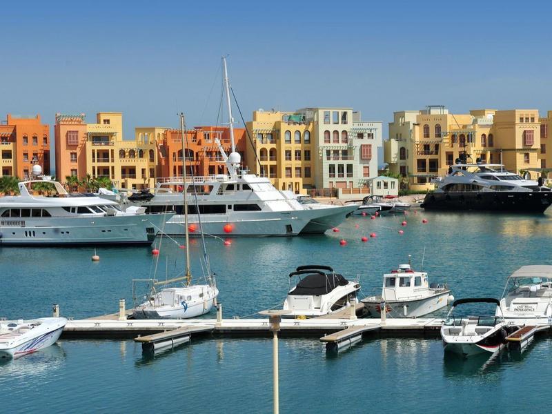 Des bâtiments colorés longent une marina avec des yachts et de petits bateaux amarrés dans une eau calme sous un ciel clair.
