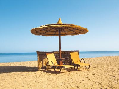 Twee ligstoelen onder een strooien parasol op een rustige zandstrand met uitzicht op zee.