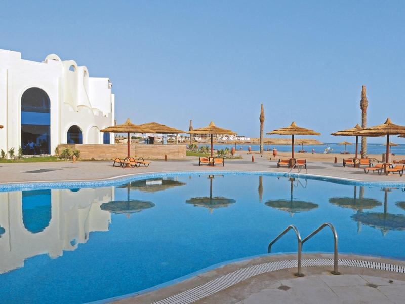 Piscina dell'hotel con ombrelloni e edifici bianchi sotto un cielo blu limpido.