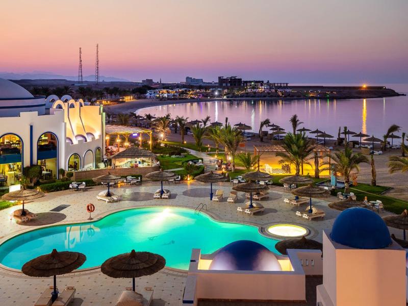 Vista serale di un hotel con piscina illuminata, palme e baia tranquilla sullo sfondo.