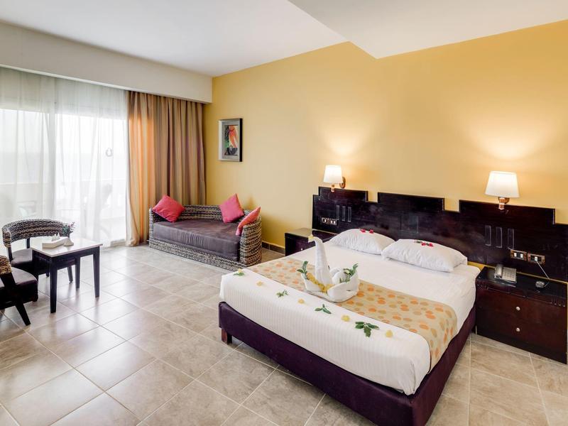 Camera d'hotel luminosa arredata con letto matrimoniale, divano, tavolo e grandi finestre.