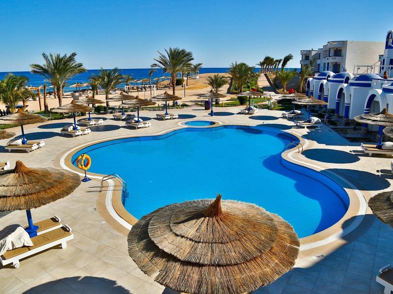 Una grande piscina a forma di goccia con lettini, ombrelloni e palme vicino al mare.