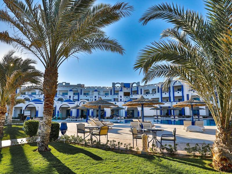 Vista di un hotel con palme e ombrelloni in primo piano sotto un cielo sereno.