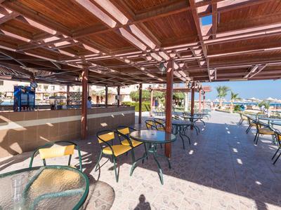 Covered terrace with tables and chairs, overlooking a pool and palm trees in sunlight.