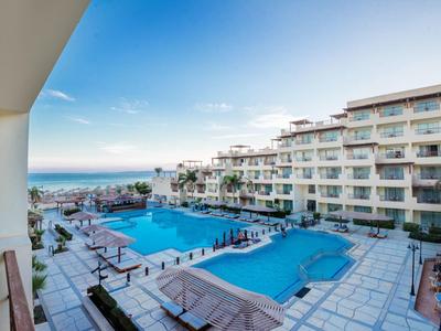 Großer Hotelpool mit Liegen, angrenzendem Gebäude und Blick auf das Meer bei blauem Himmel.