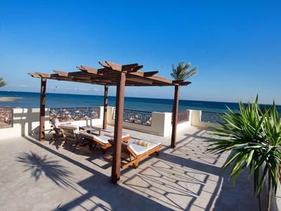 Beach terrace with wooden pergola, loungers, and view of the blue sea under clear sky.