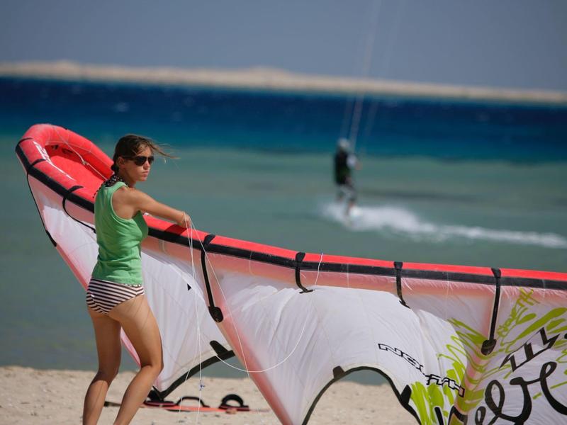 Vrouw op strand met windsurfzeil voor turquoise zee en kitesurfer op de achtergrond.