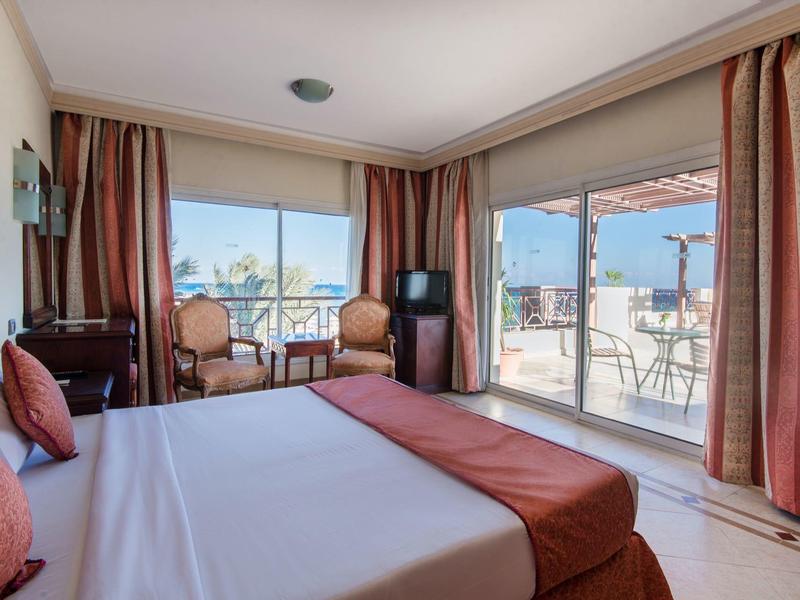 Hotelkamer met groot bed, twee fauteuils, tv en toegang tot balkon met zeezicht.