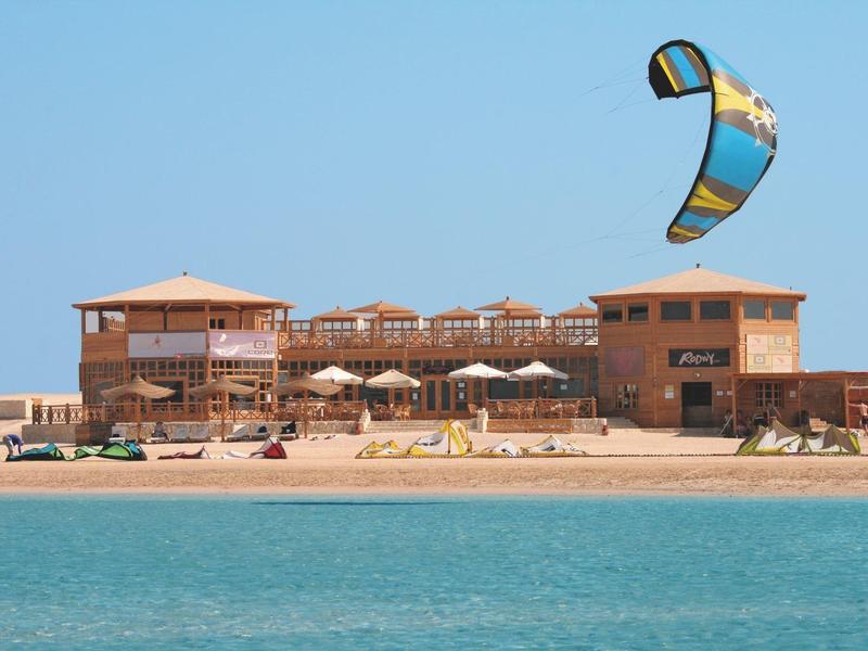 Strand mit gelbem Sand, blauem Meer, Kitesurfer und großem Holzgebäude im Hintergrund.