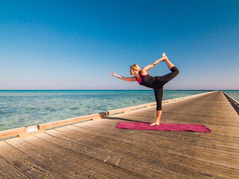Frau macht Yoga auf roter Matte am Holzsteg mit Meer und blauem Himmel im Hintergrund.