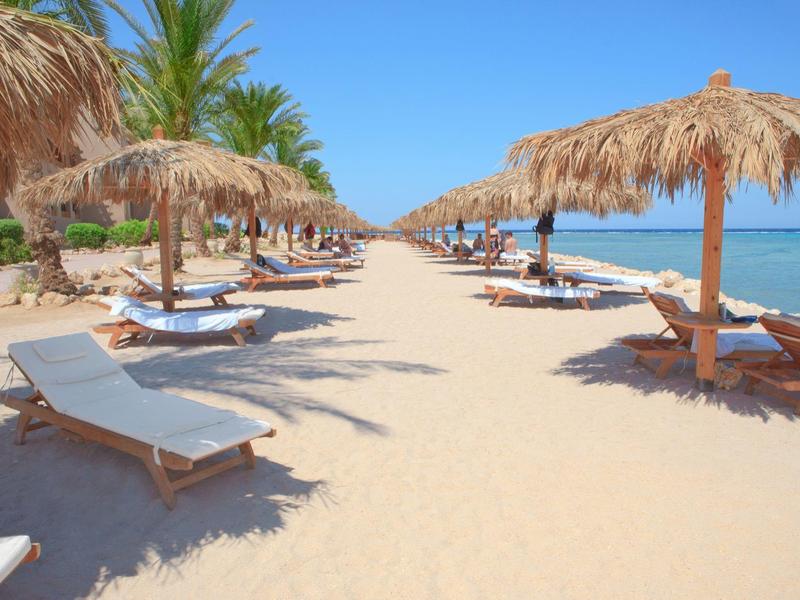Strand mit weißen Liegestühlen und Strohm Sonnenschirmen, blauer Himmel und Palmen im Hintergrund.