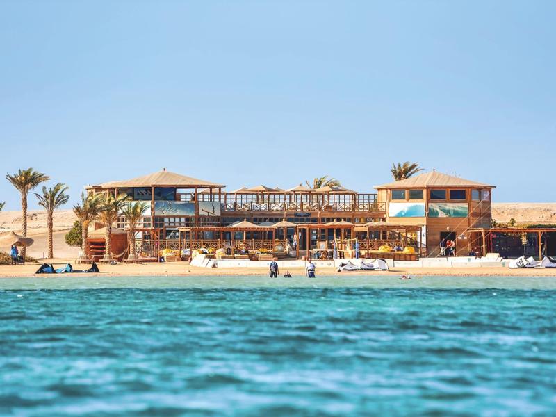 Strandrestaurant mit Holzterrassen, Palmen und blauer See unter klarem Himmel.