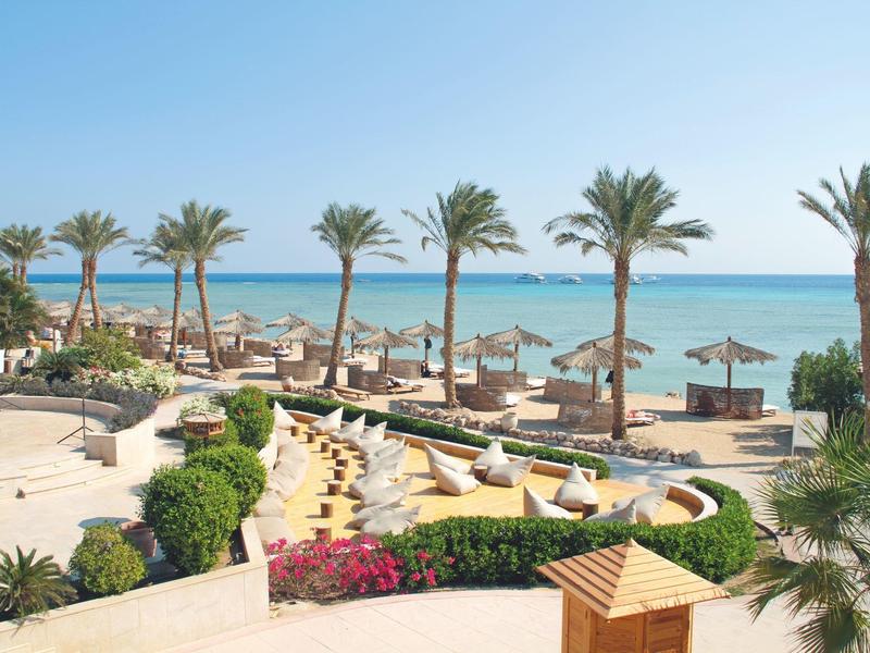 Strand mit Palmen, Sonnenschirmen und blühenden Pflanzen unter klarem blauem Himmel.