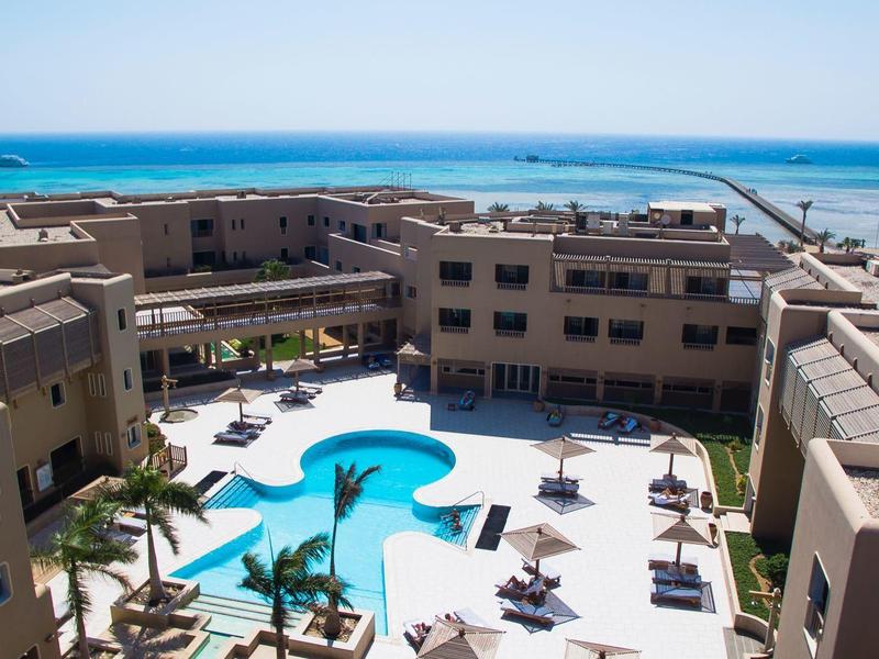 Hotel mit rundem Pool, Sonnenschirmen und Blick auf das blaue Meer im Hintergrund.