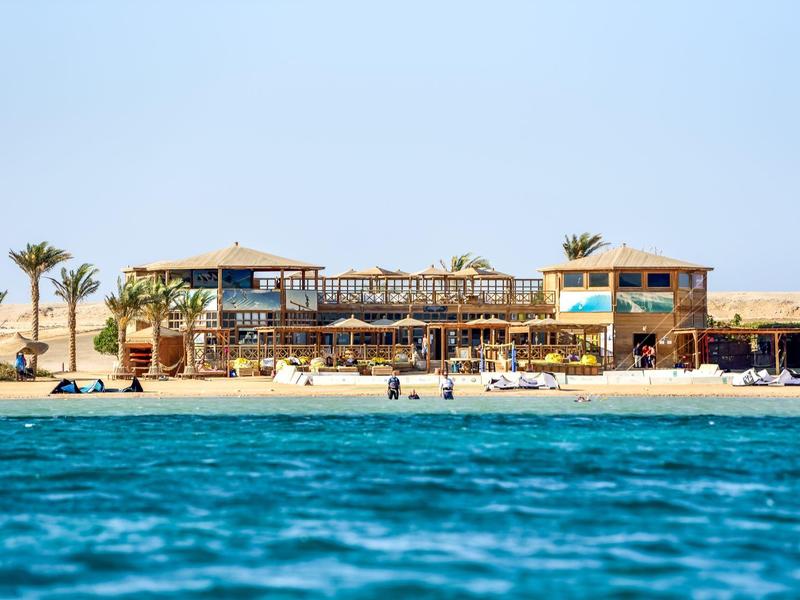 Strand mit blauem Meer, Palmen und mehreren flachen Gebäuden unter klarem Himmel.