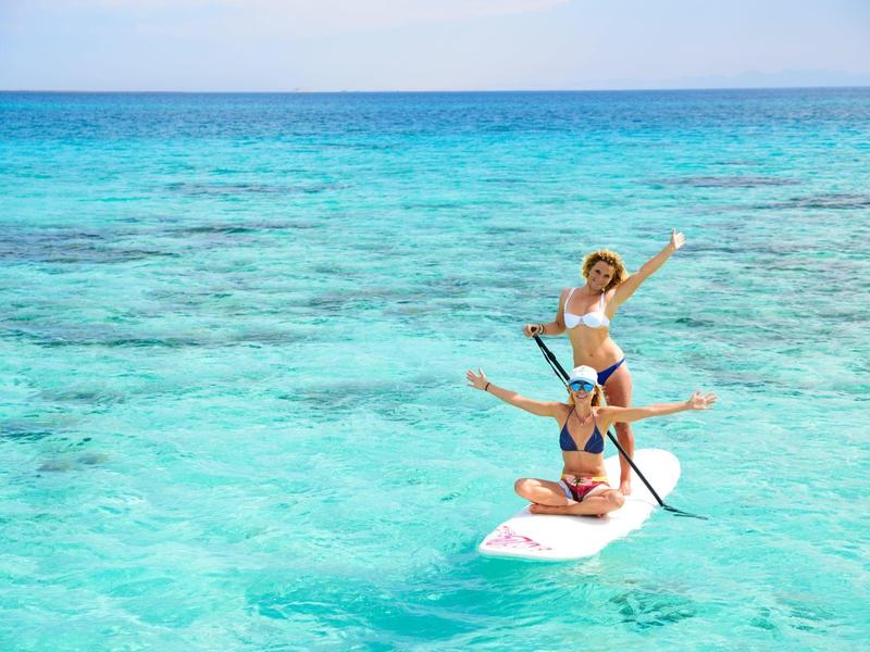 Zwei Frauen auf Stand-Up-Paddle-Board im klaren, türkisfarbenen Meer bei sonnigem Himmel.