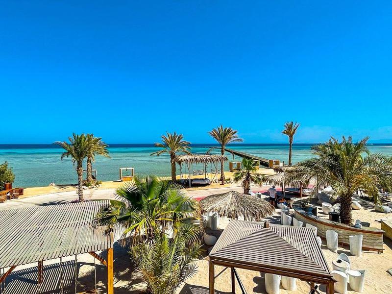 Strand mit Palmen, Holzterrassen, Sitzbereichen und klarem blauem Himmel über ruhigem Meer.