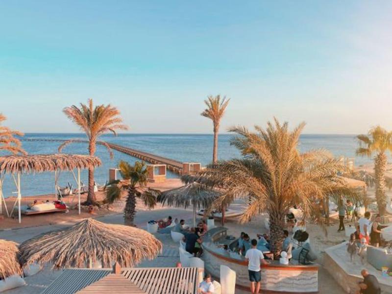 Strandbar mit Palmen, Strohdächern und Sitzbereichen an einem sonnigen, blauen Meereshorizont.