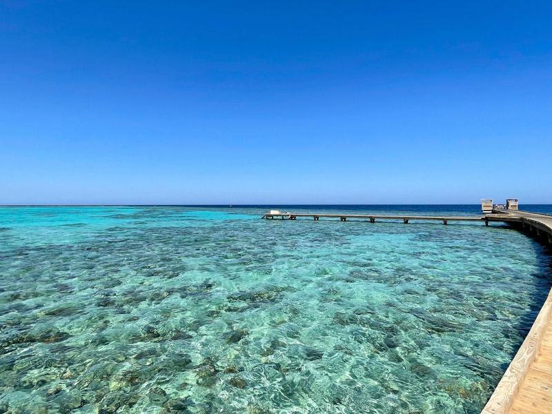 Klares türkisfarbenes Meer mit Holzsteg, der sich ins Wasser hinauskrümmt unter blauem Himmel.