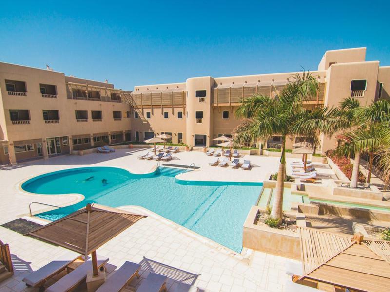 Innenhof eines Hotels mit Schwimmbad, Sonnenterrasse und Palmen unter blauem Himmel.