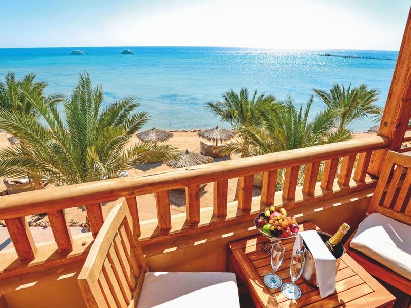Balkon mit Holzgeländer und Stühlen mit Meerblick und Palmen am Strand.