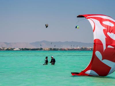 Due subacquei stanno in acqua turchese vicino a una costa con montagne e una vela da kitesurf colorata in primo piano.