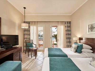 Hotelkamer met twee bedden, teal accenten, tv en balkondeuren die daglicht binnenlaten.