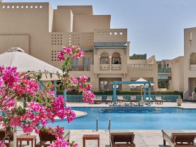Una piscina all'aperto di un hotel con fiori rosa in fiore in primo piano e edifici beige sullo sfondo.