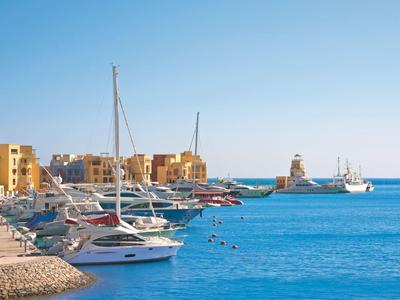 Jachten aangemeerd in een jachthaven met gebouwen en een vuurtoren op de achtergrond onder een heldere hemel.