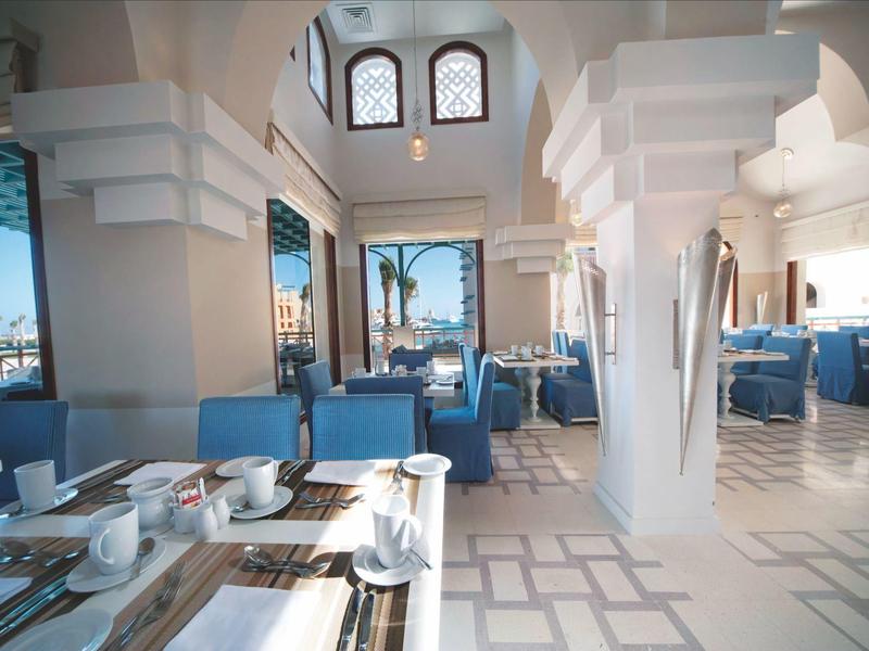 Zonnige hotel eetruimte met witte bogen, blauwe stoelen en uitzicht op de oceaan.