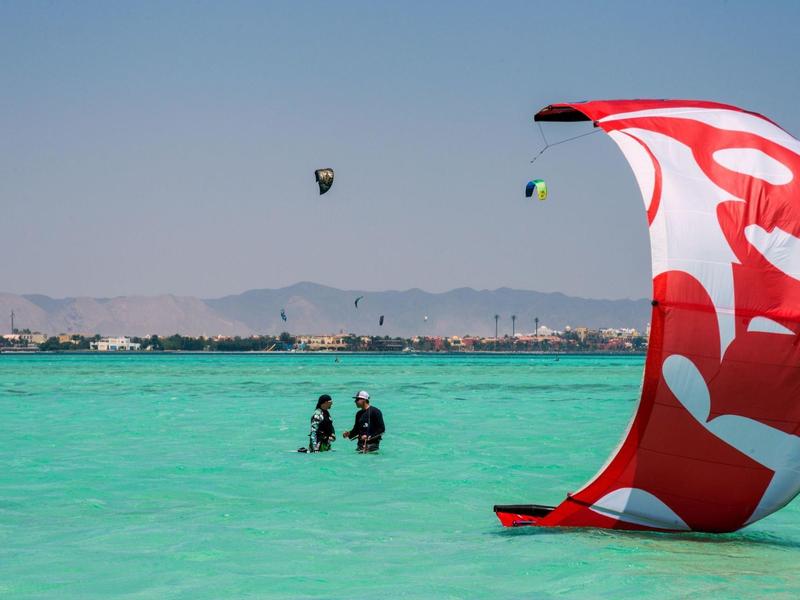 Strand met turquoise water, kitesurfers en een rood-witte vlieger op de voorgrond.