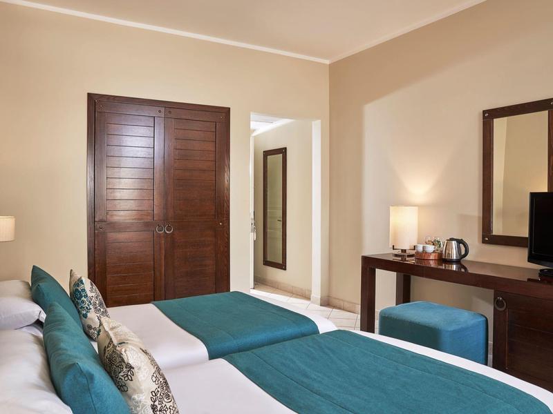 Hotelkamer met twee bedden, teal bedlopers, houten kast, tv en spiegel aan beige muren.