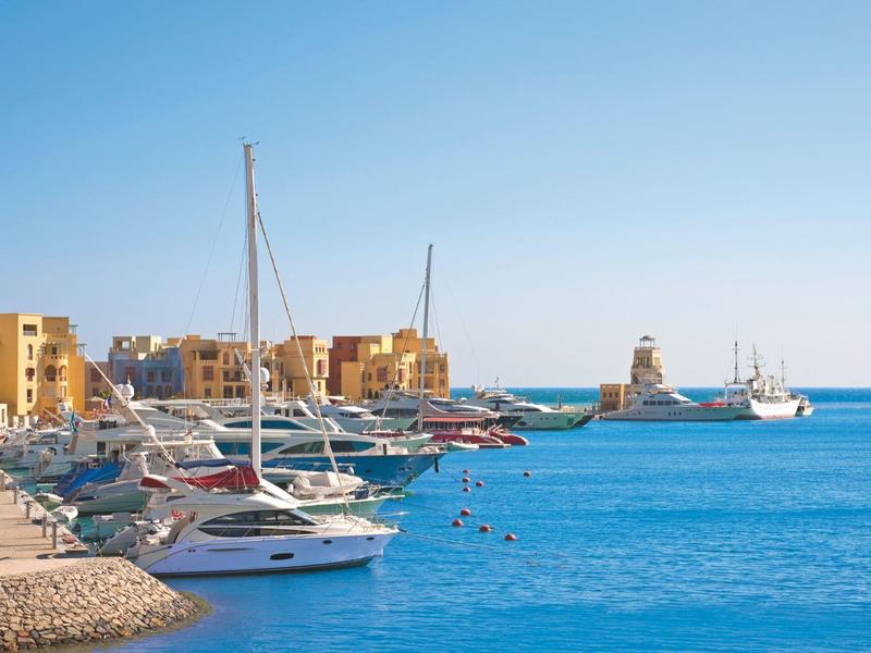 Jachten aangemeerd in een jachthaven met gebouwen en een vuurtoren op de achtergrond onder een heldere hemel.