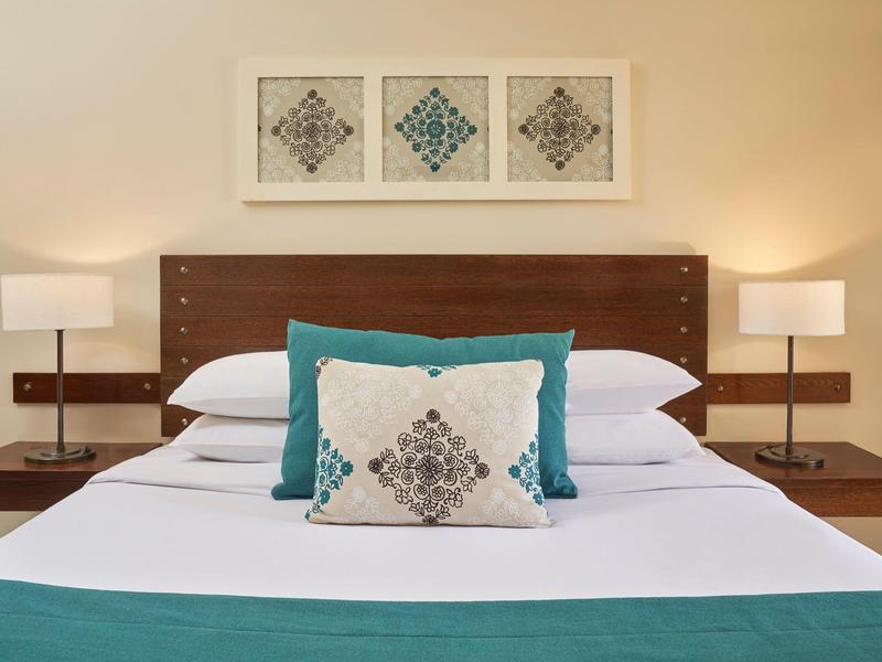 Gezellig hotelbed met houten hoofdeinde, teal en patroon kussens, en nachtlampjes.