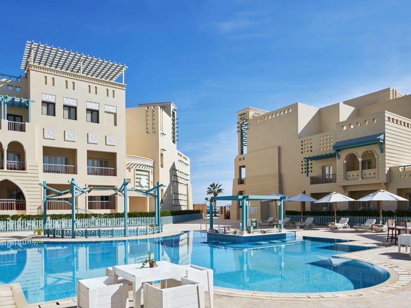 Buitenzwembad met ligstoelen en tafels bij een zonnig resort onder een heldere blauwe lucht.