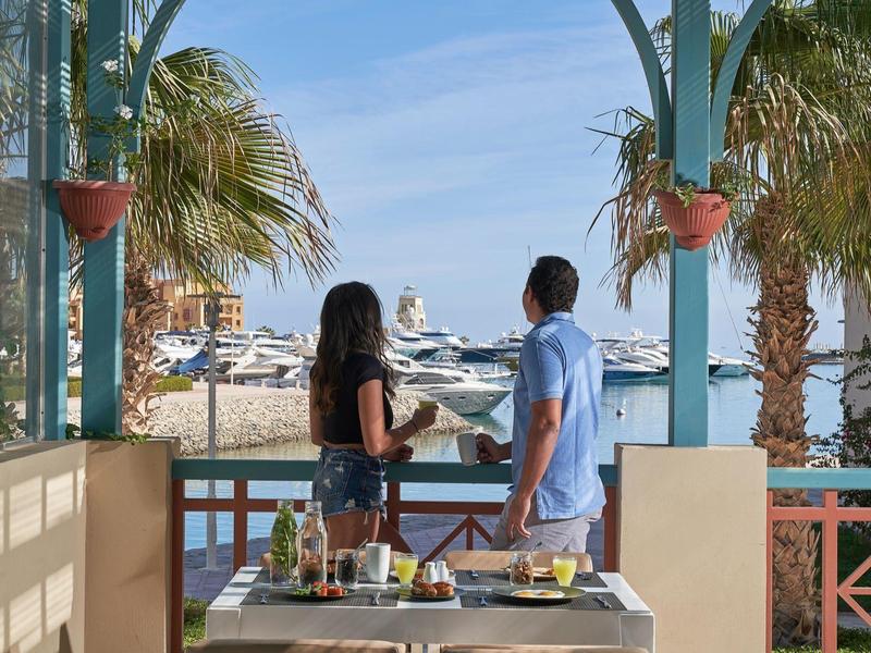 Stel geniet uitzicht op een jachthaven vanaf een terras met een eettafel en palmbomen.