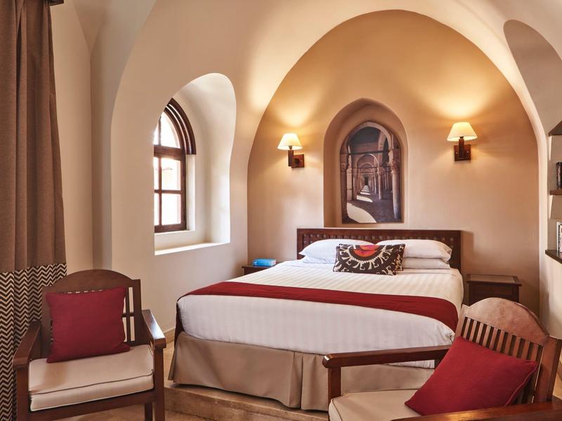 Chambre d'hôtel confortable avec fenêtres en arc, lit double, fauteuils et éclairage chaleureux.