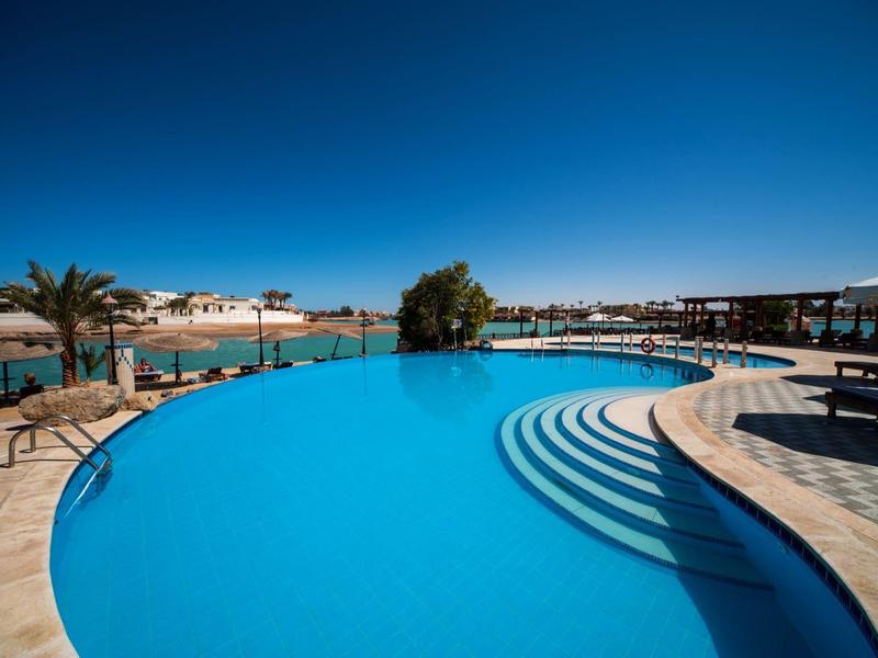 Piscine à débordement incurvée avec vue sur l'eau, palmiers et transats sous un ciel bleu clair.